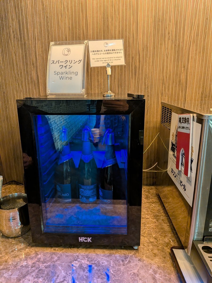 青い光でライトアップされた小型冷蔵庫に、数本のスパークリングワインボトルが冷やされている様子。上には「スパークリングワイン」の案内表示と、飲酒に関する注意書きが置かれている。