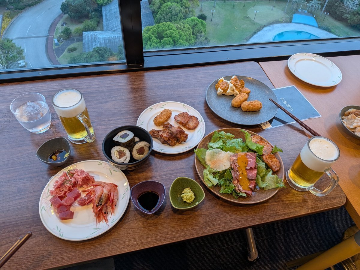 窓際のテーブルに、ビールジョッキや水、刺身、巻き寿司、ローストビーフサラダ、揚げ物など、バラエティ豊かな料理が並べられている。窓の外には夜景が広がっている。