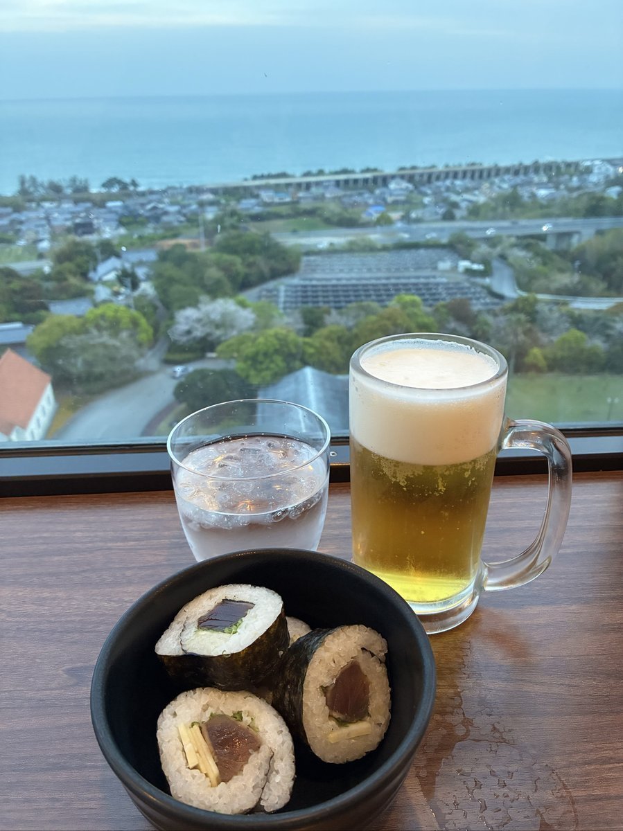 窓際のテーブルに置かれたビールジョッキと巻き寿司の小鉢。窓の外には、広がる海と遠くの街並みが広がる開放的な景色が見える。