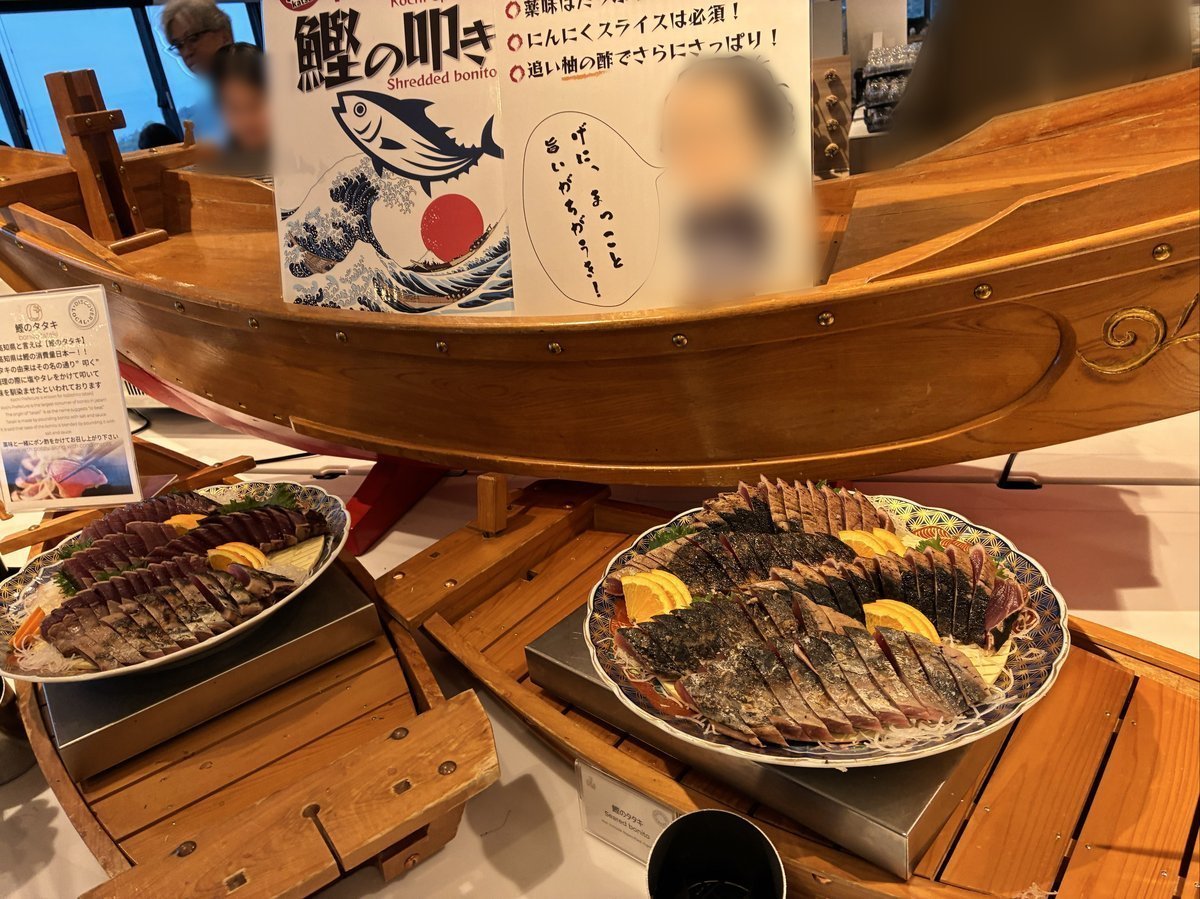 木製の船形ディスプレイに、大量の鰹のたたきが盛り付けられた大皿が二つ並べられている。上には「鰹のたたき」と書かれた大きな看板がある。