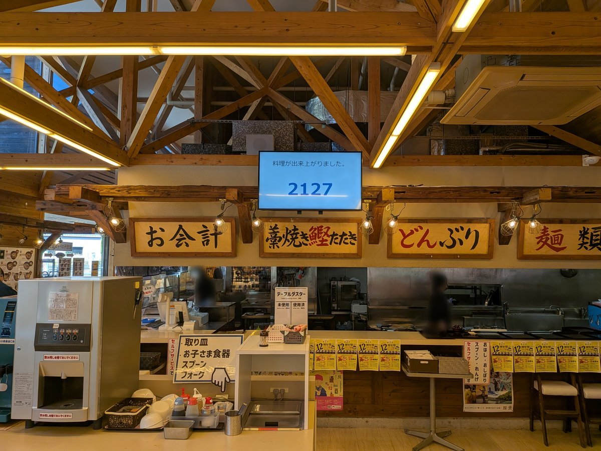 木の温もりを感じるフードコートの注文カウンター。番号表示モニターの下には「お会計」「藁焼きたたき」などの看板が並び、活気ある雰囲気。セルフサービスコーナーも完備。