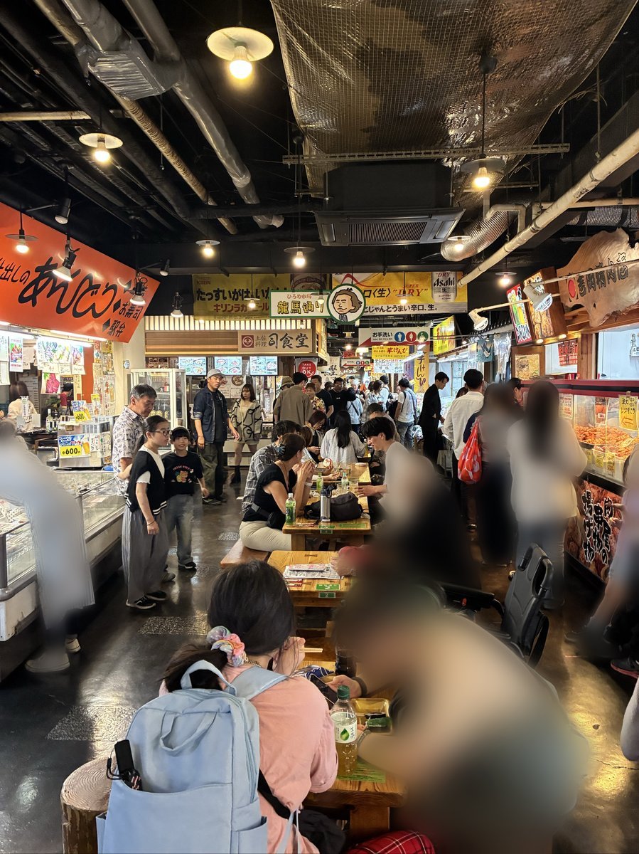 市場内部の活気ある様子。多くの人々がテーブル席で食事をしたり、通路を行き交ったりしており、賑やかな雰囲気が伝わる。天井には配管や照明が見える。