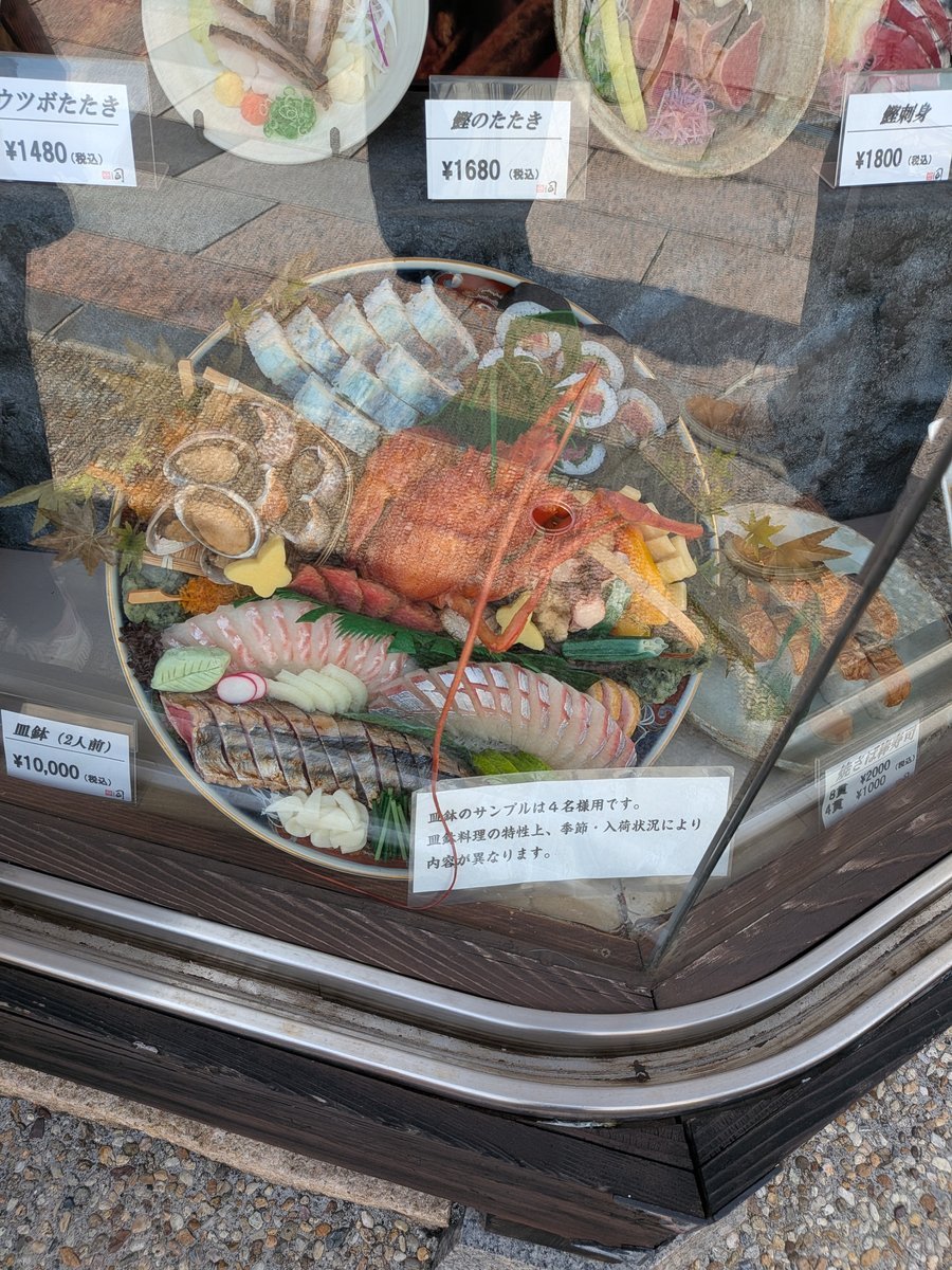 店舗のショーケースに飾られた、豪華な皿鉢料理の食品サンプル。大きな伊勢海老のような食材を中心に、刺身や寿司、煮物などが美しく盛り付けられている。周辺には他の海鮮料理のサンプルと価格表示が見える。