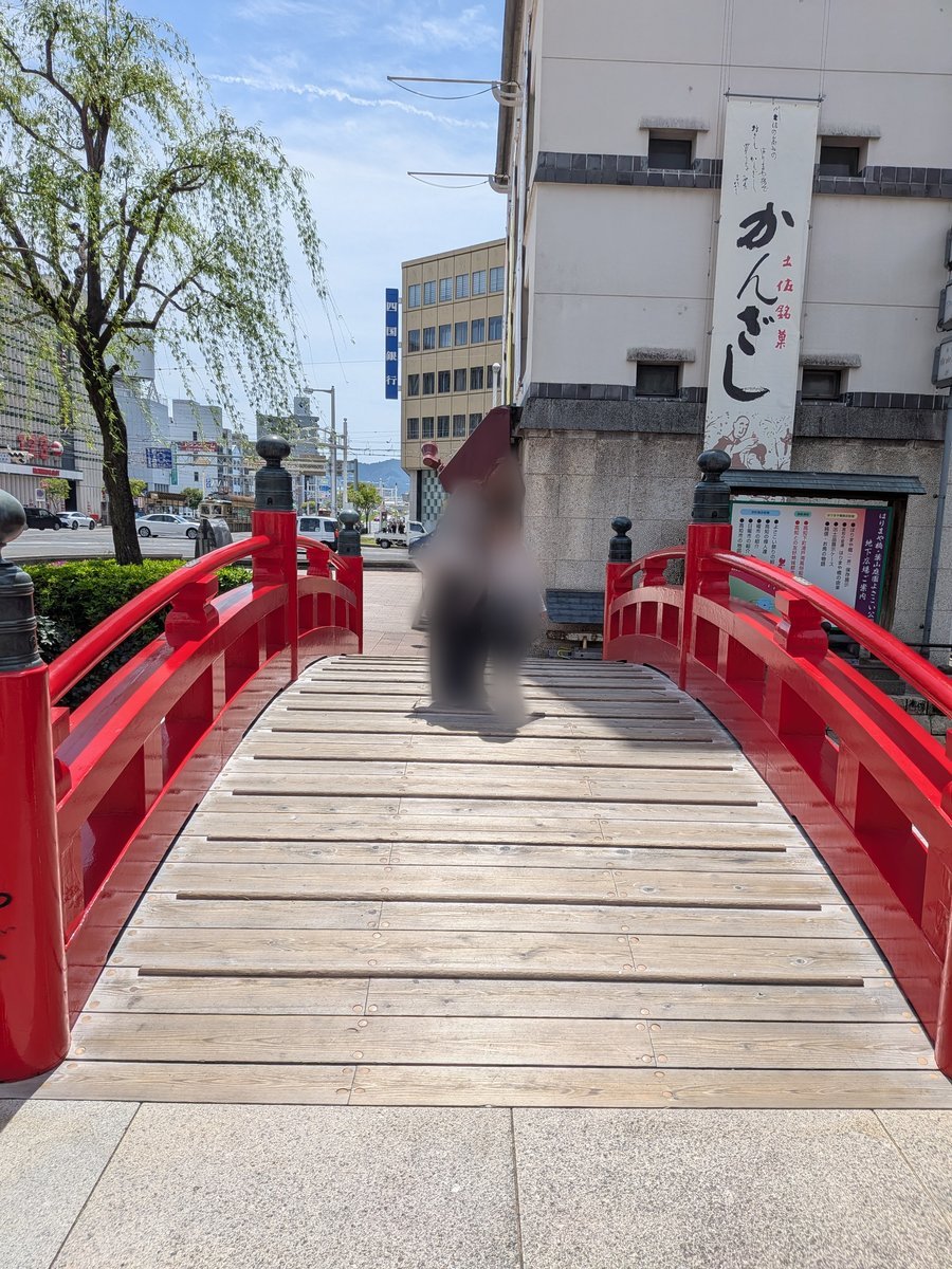 朱色の欄干が特徴的な小さな太鼓橋。木製の橋板が敷かれ、奥には街並みが広がる。橋の向こうの建物には「かんざし」の看板が見える。