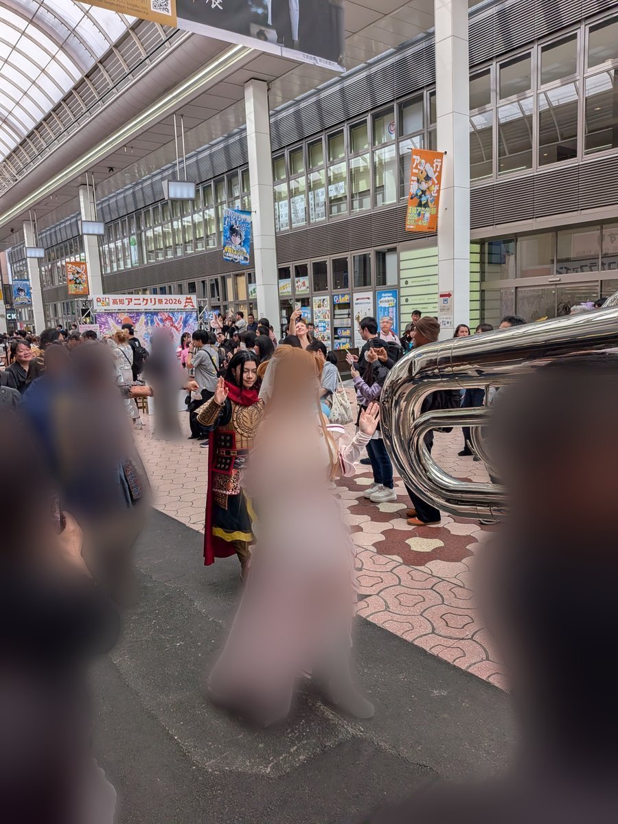 屋根のある商店街で、多くの人々で賑わうイベントの様子。様々なコスチュームを身につけた参加者が見られ、大きな金管楽器も写っている。活気ある「高知アニクリ祭」の雰囲気が伝わる。