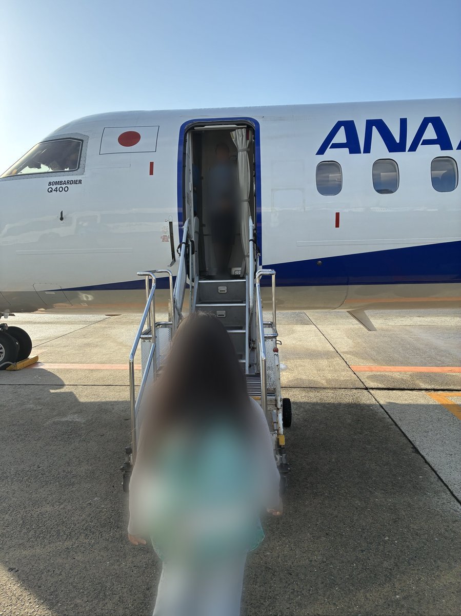 青い空の下、ANAのプロペラ機「ボンバルディアQ400」にタラップを使って搭乗する様子。機体には日の丸が描かれている。