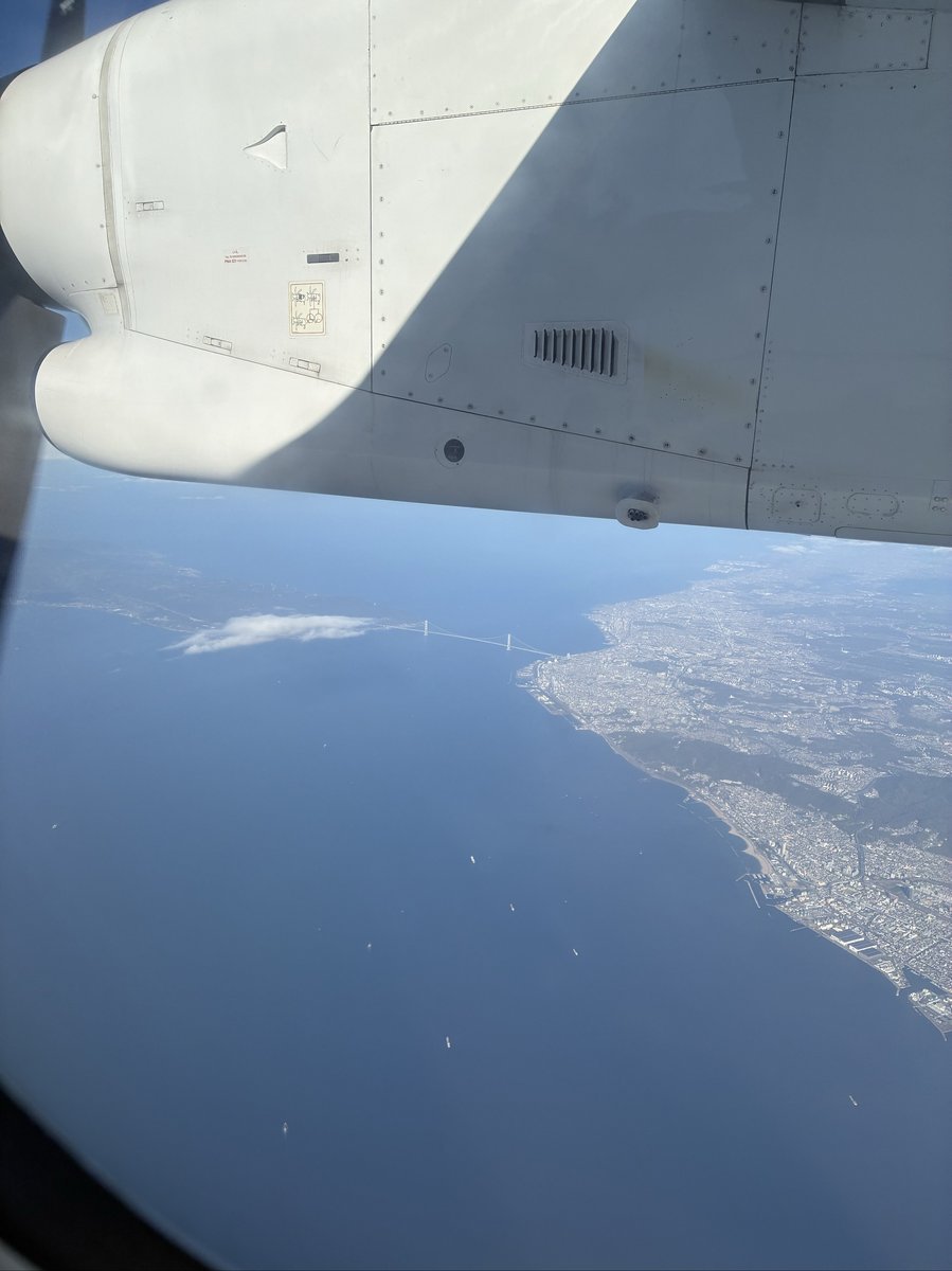 飛行機の窓から見下ろす、広大な海と陸地を結ぶ明石海峡大橋。遠くには街並みが広がり、雄大な景色が広がっている。