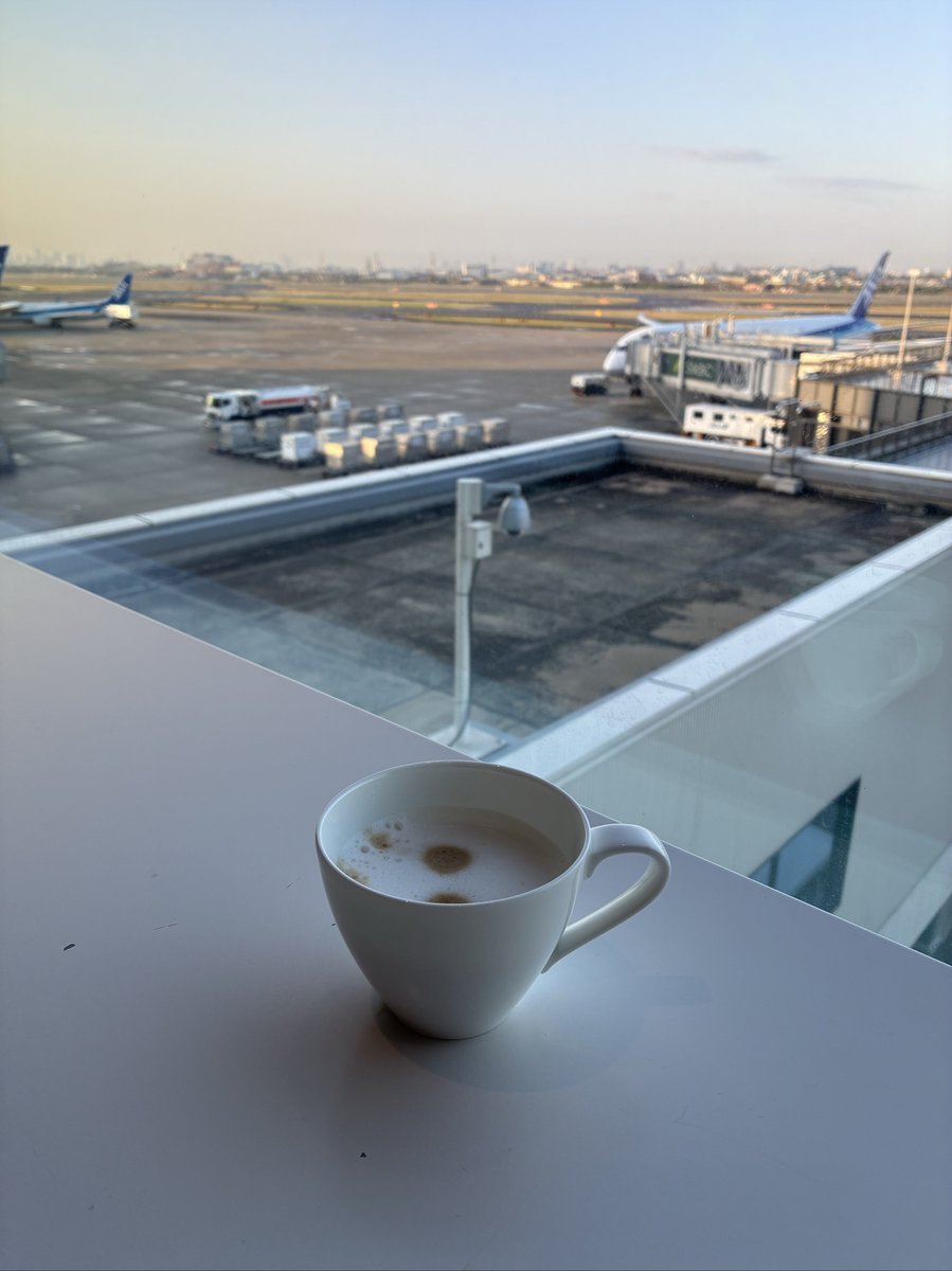 空港ラウンジの窓際に置かれたカフェラテのカップ。窓の外には離発着する飛行機や滑走路が広がり、旅情を誘う。
