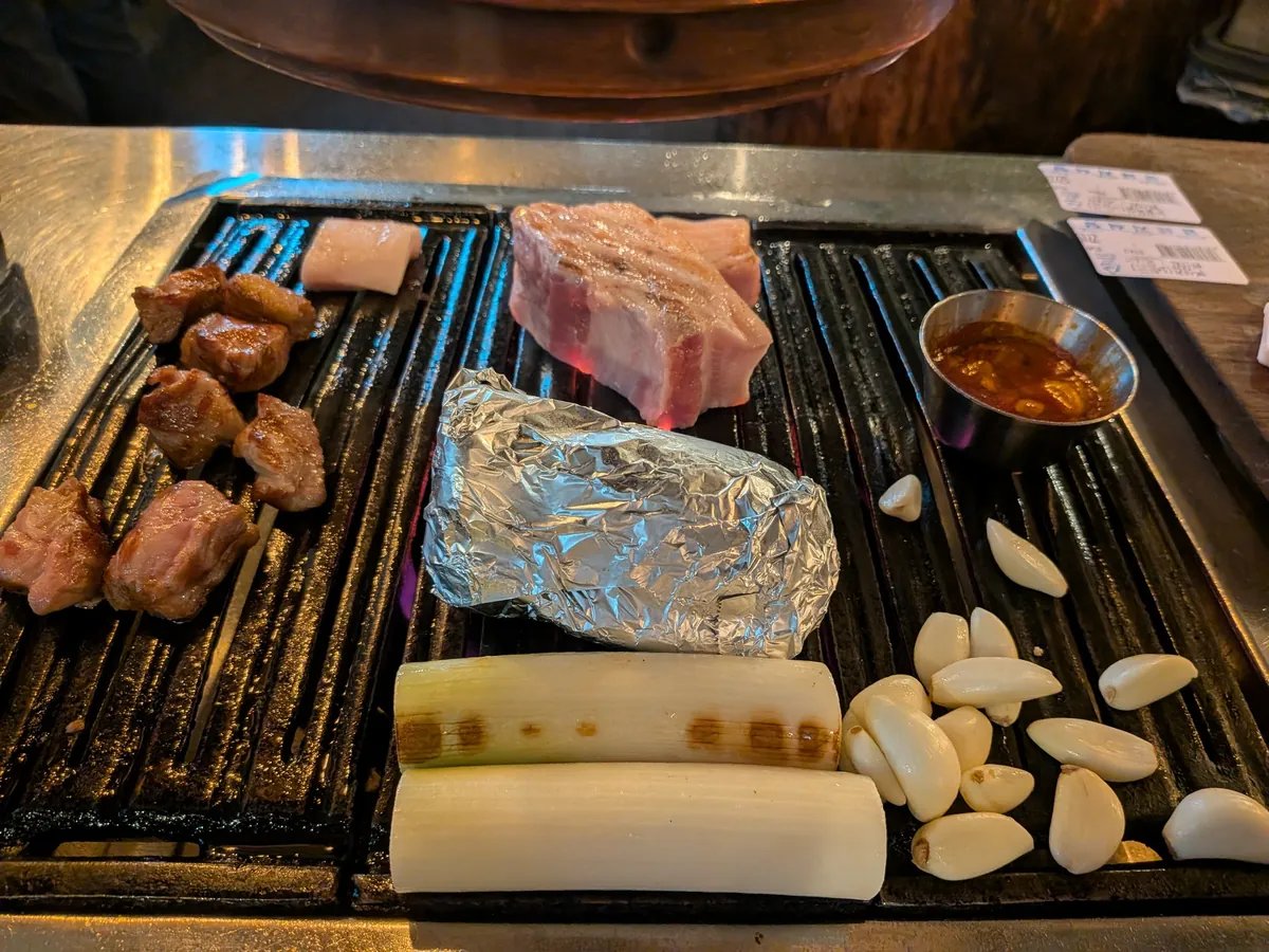 熱された焼肉用鉄板にサムギョプサルの厚切り豚肉や一口大の肉、白い脂身が並べられている。隣にはアルミホイルに包まれたもの、焦げ目のついた長ネギ、そして大量の剥きニンニクが置かれ、焼かれている最中である。肉を食べるためのタレが入った小さな器も見える。