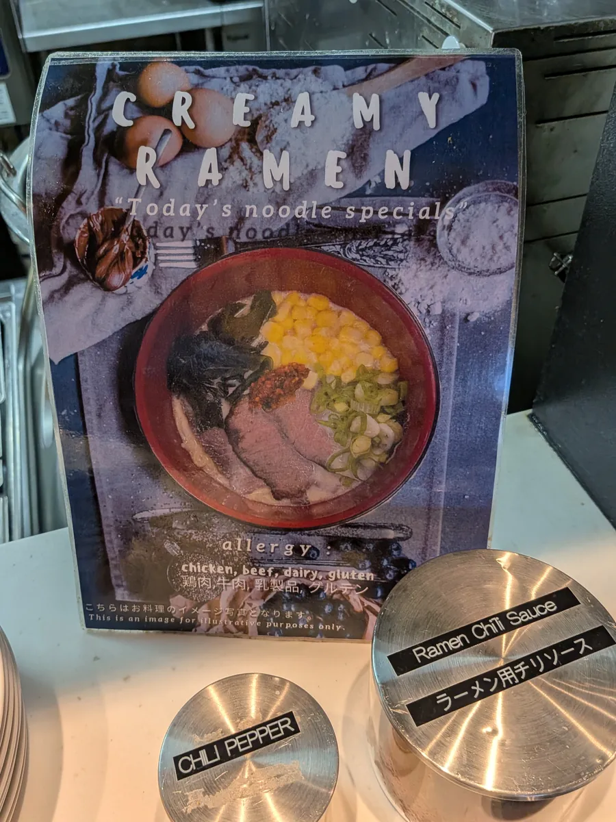 ラウンジのヌードルコーナーに置かれた「CREAMY RAMEN」の掲示。本日のヌードルスペシャルとして紹介され、隣にはラーメン用チリソースとチリペッパーの容器が並ぶ。