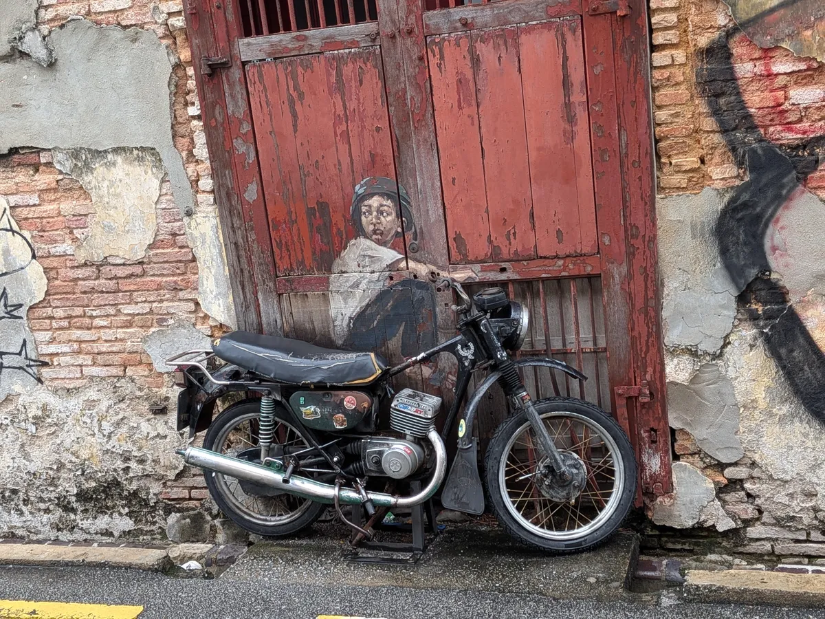 アークィー通りの壁画Boy on Motorbike。赤い木の扉の前に本物の古いバイクが置かれ、壁に描かれた少年と一体になっている