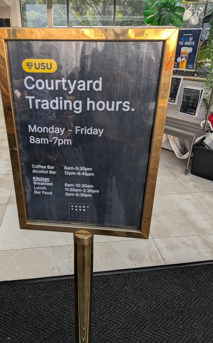 Courtyardの営業時間を示す金縁の立て看板。月曜から金曜8時から19時、コーヒーバー・アルコールバー・キッチンの時間帯が書かれている