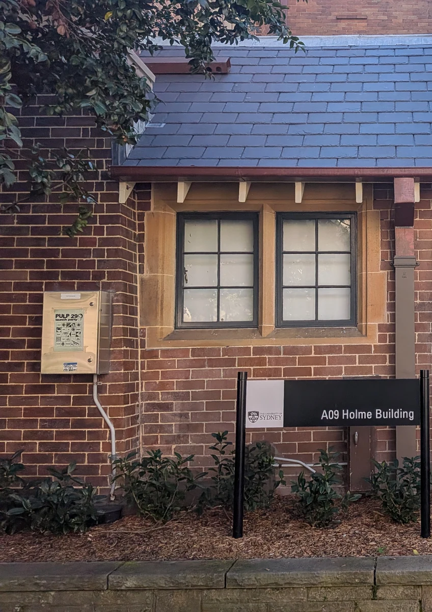 シドニー大学のA09 Holme Buildingの外観。レンガ造りの壁と青いスレート屋根、格子窓の下に建物名の表示板が立っている