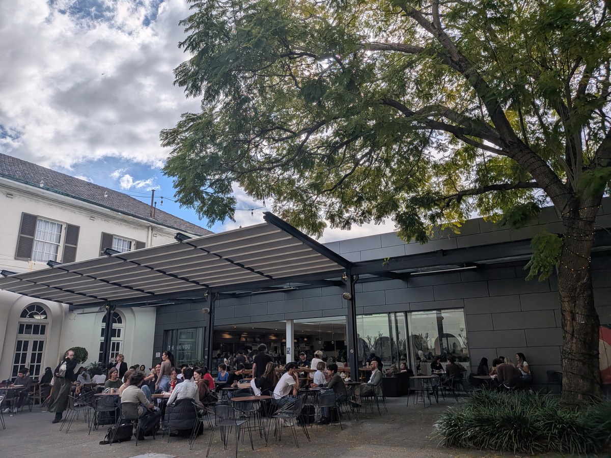シドニー大学Courtyard Restaurant & Barの屋外テラス席。大きな木の下に白い日よけが張り出し、学生や教員でほぼ満席になっている