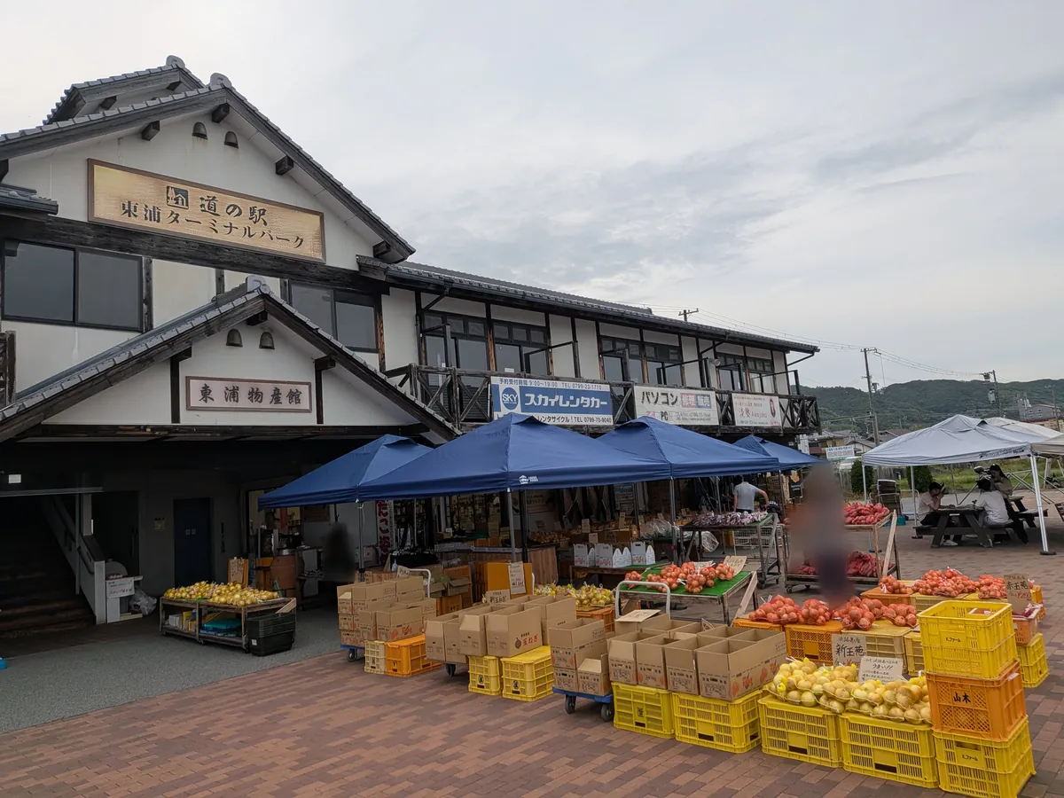 道の駅 東浦ターミナルパークの本館外観と屋外の青いテントの野菜販売エリア、玉ねぎの入った黄色いコンテナ
