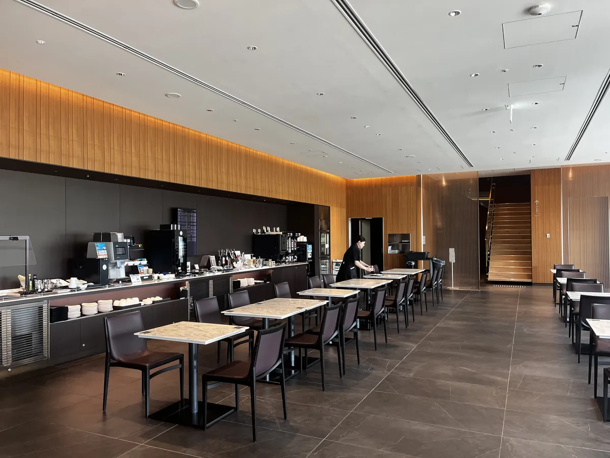 食事エリアのテーブル席とビュッフェカウンター、コーヒーマシン。奥に木の階段が見える広い空間。