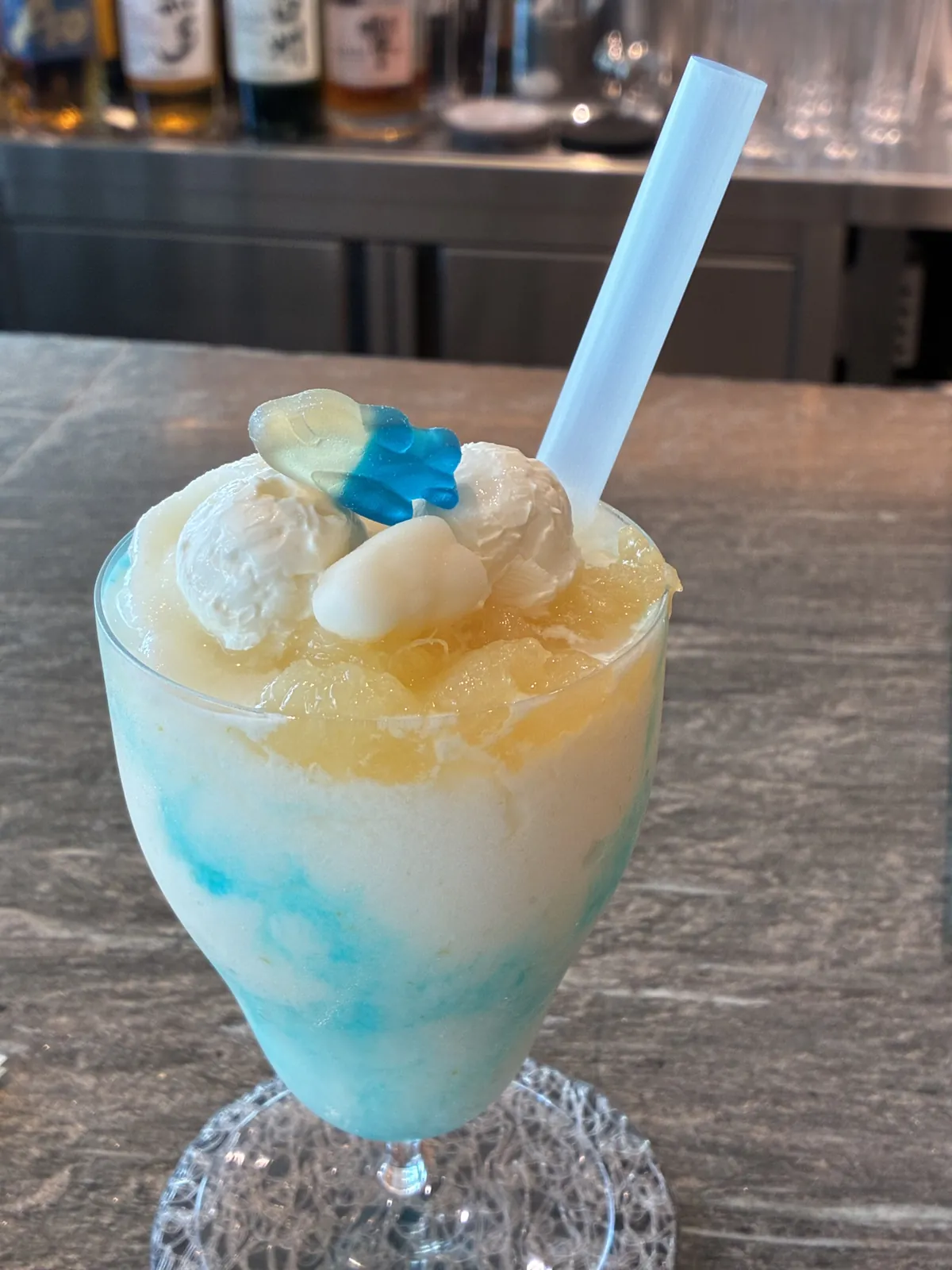ANAスイートラウンジの夏限定ノンアルフローズンモクテル(ANA Ice Magic 夏バージョン)。青×白×黄のフローズンに生クリームと青いグミ、奥はバーのボトル棚。