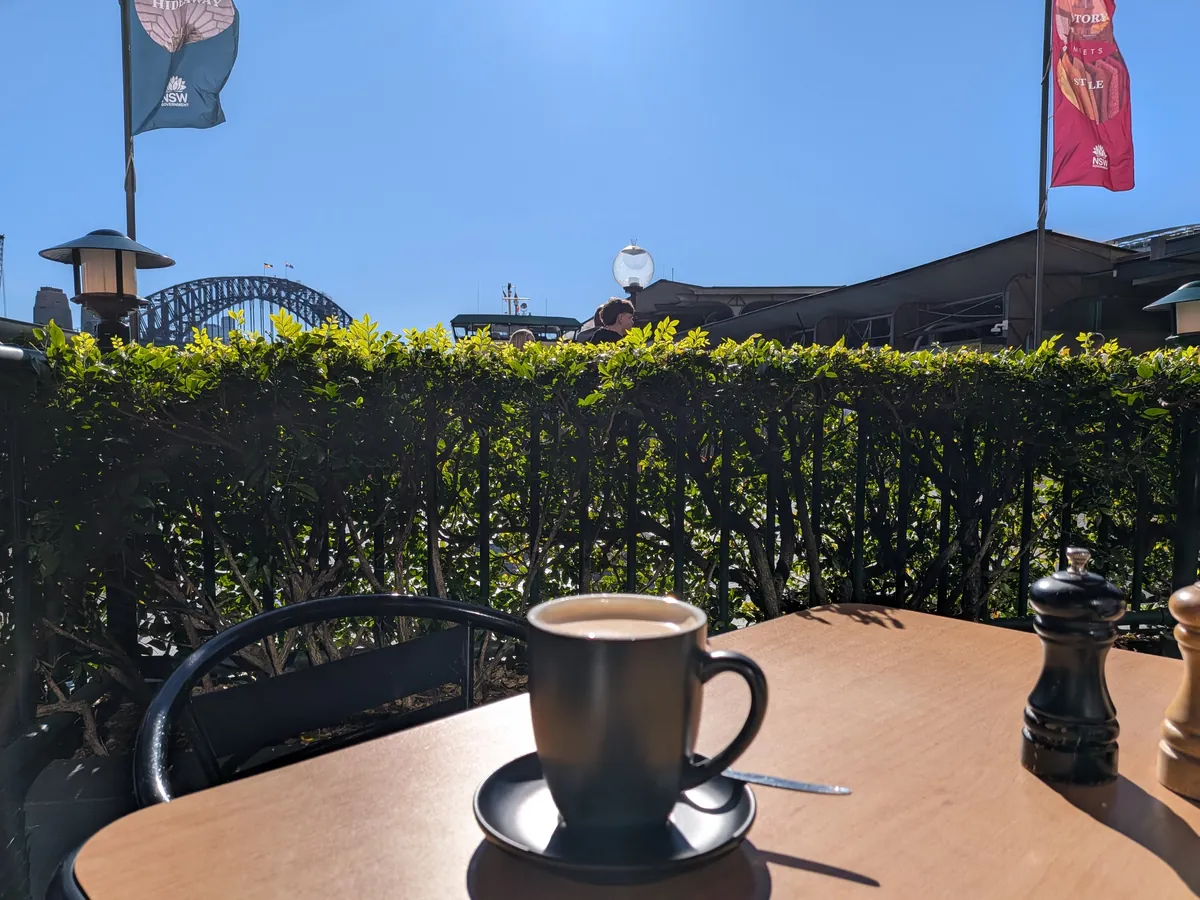 City Extraのテラス席のテーブルに置かれたコーヒーと、生垣の向こうの旗とサーキュラーキーの空