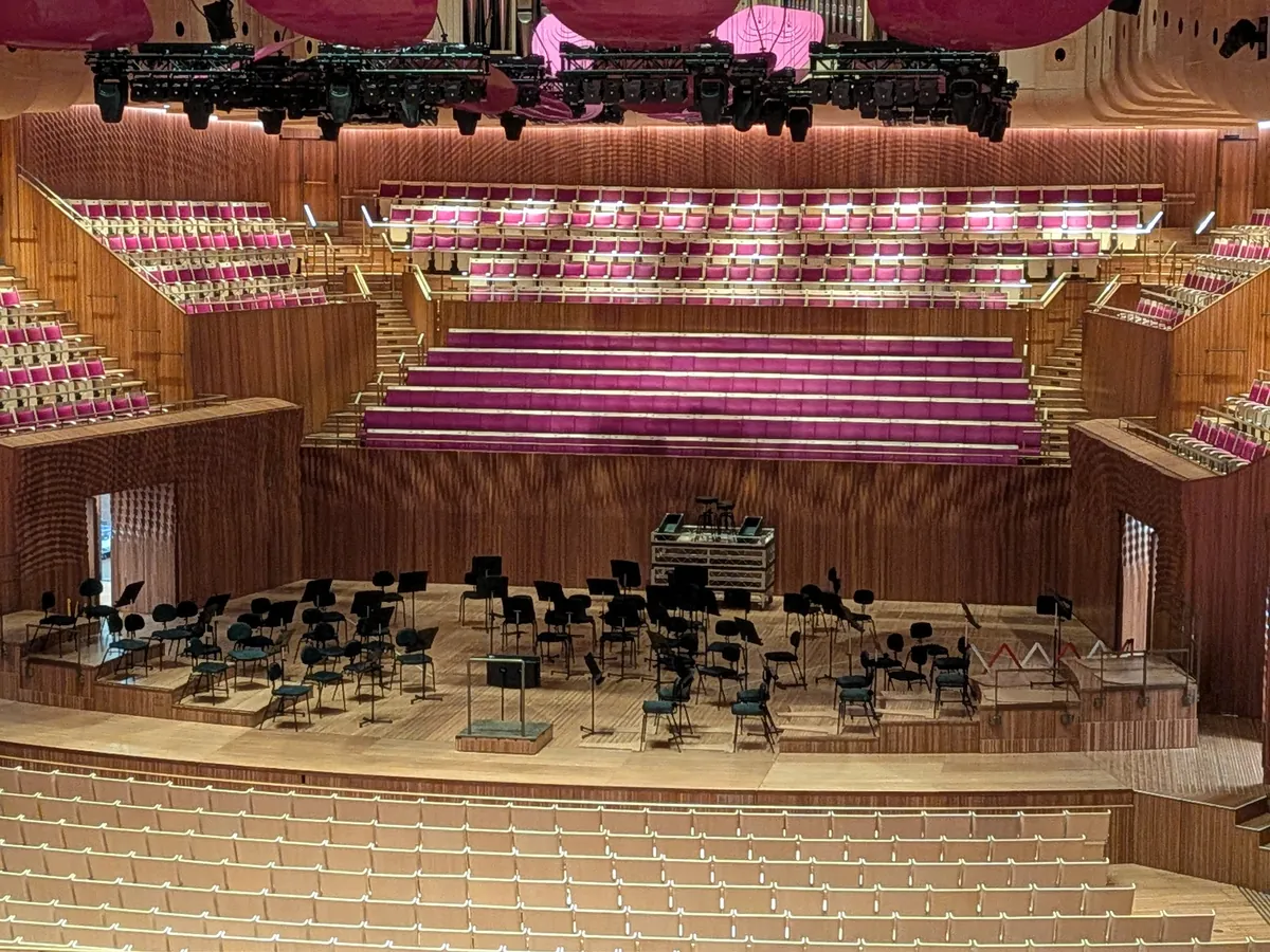 コンサートホールのステージ。公演に向けて椅子と譜面台が並べられ、背後にはピンクの客席が段状に広がる