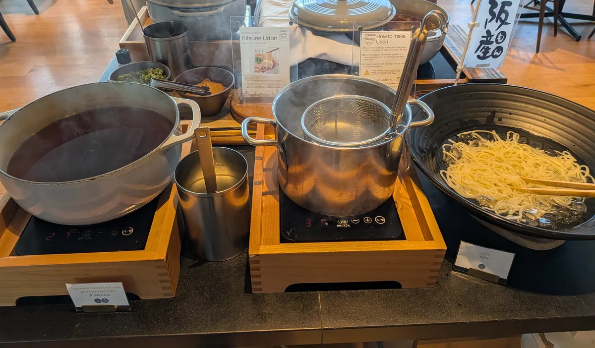 自分でうどんを茹でられるヌードルコーナー。出汁の鍋、茹で用の湯とテボ、生うどんが並ぶ