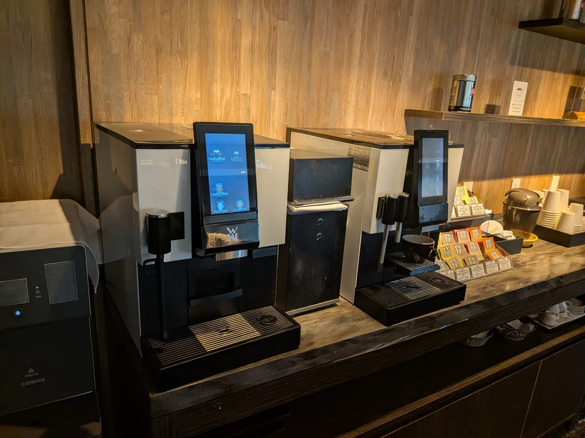 ラウンジのタッチパネル式コーヒーマシン2台とカップが並ぶドリンクコーナー