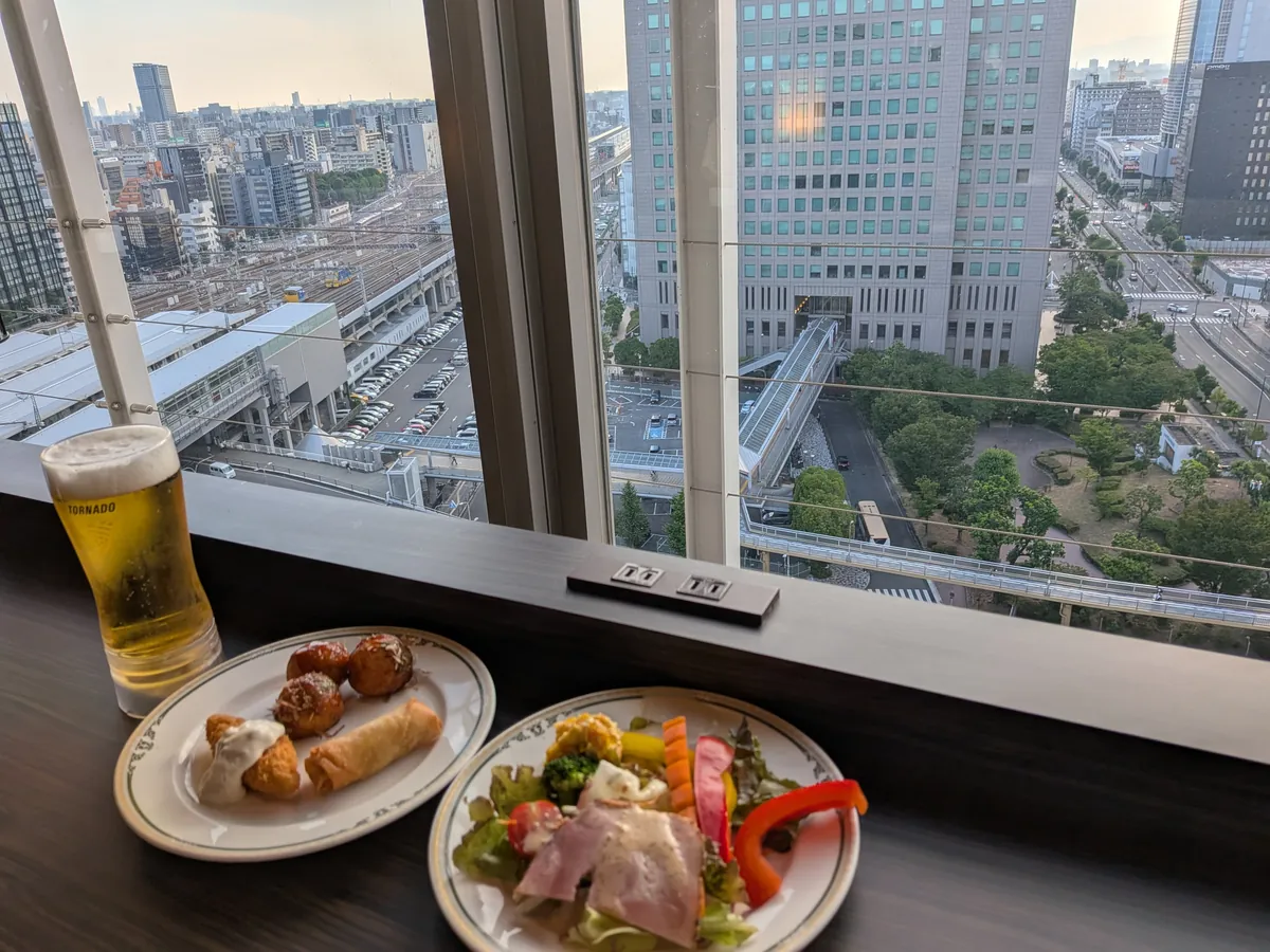 窓際の席に置かれた生ビールとオードブルの皿。夕暮れの新大阪駅を見下ろす