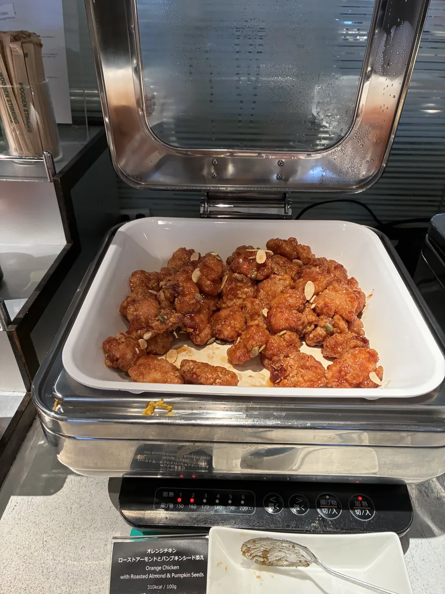 ラウンジの温かい料理コーナーに並べられた、ローストアーモンドとパンプキンシードが添えられたオレンジチキン。