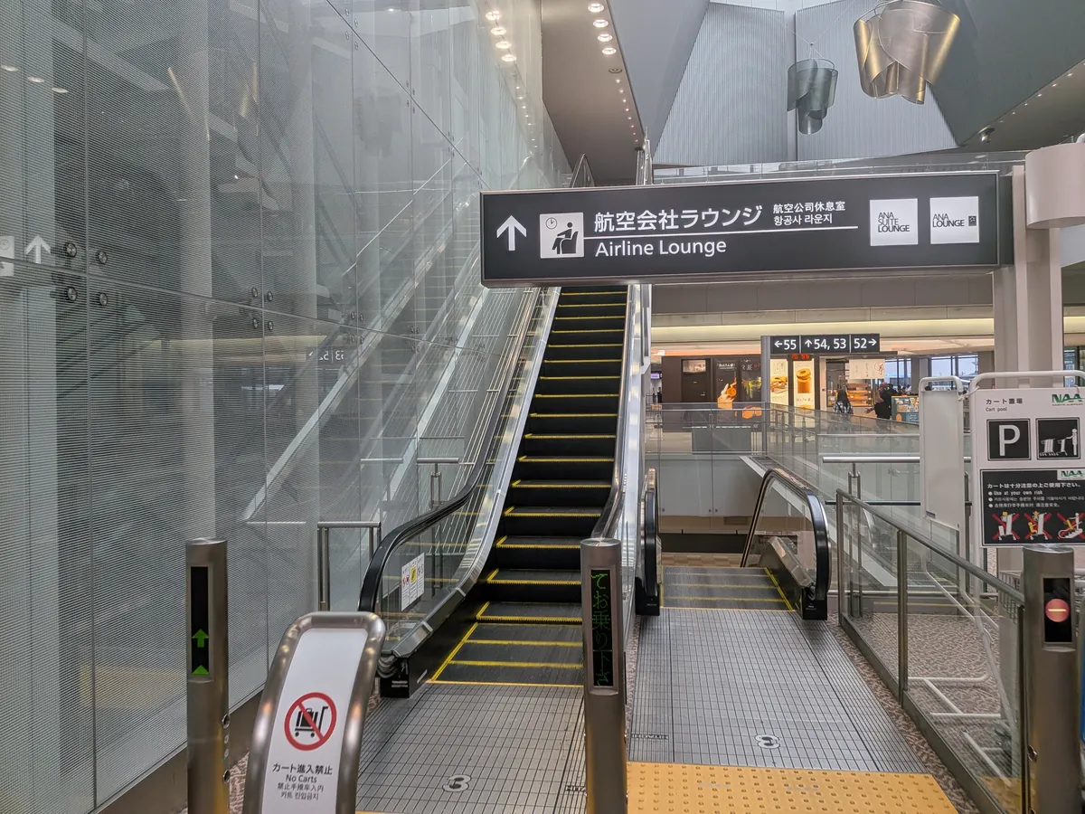 空港ターミナル内のエスカレーターと、上階にある航空会社ラウンジへの案内表示。手前にはカート禁止のマークがある。