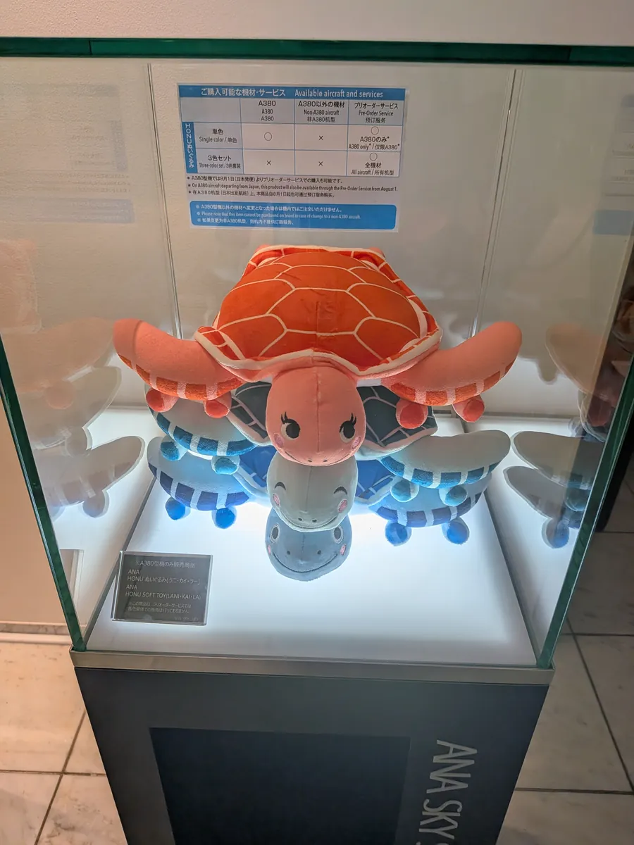 ANA SKY SHOPの展示ケースに飾られた、フライングホヌのキャラクター（オレンジのラーと青いラニ）のぬいぐるみ。