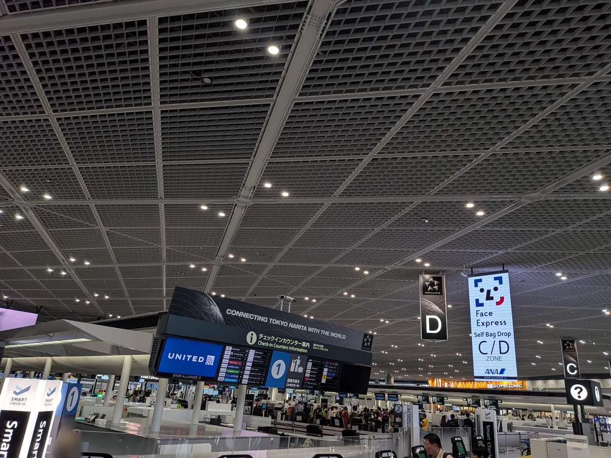 空港ターミナル内のチェックインカウンター付近の様子。格子状の天井と、国際線への接続や顔認証システム「Face Express」を案内する大きな表示板が特徴的。