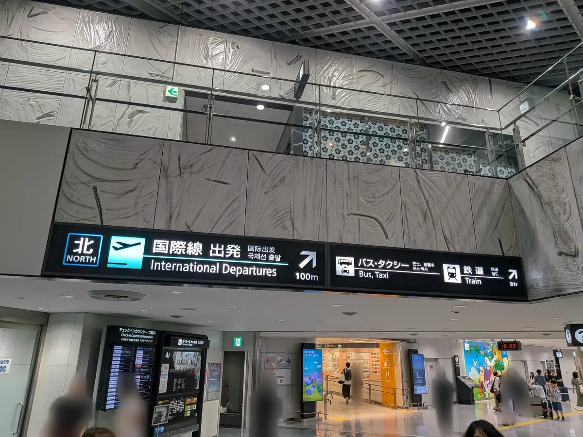 空港ターミナル内の案内表示板。「国際線 出発」や「バス・タクシー」「鉄道」といった交通機関への誘導が示されており、壁面には特徴的な模様が施されている。