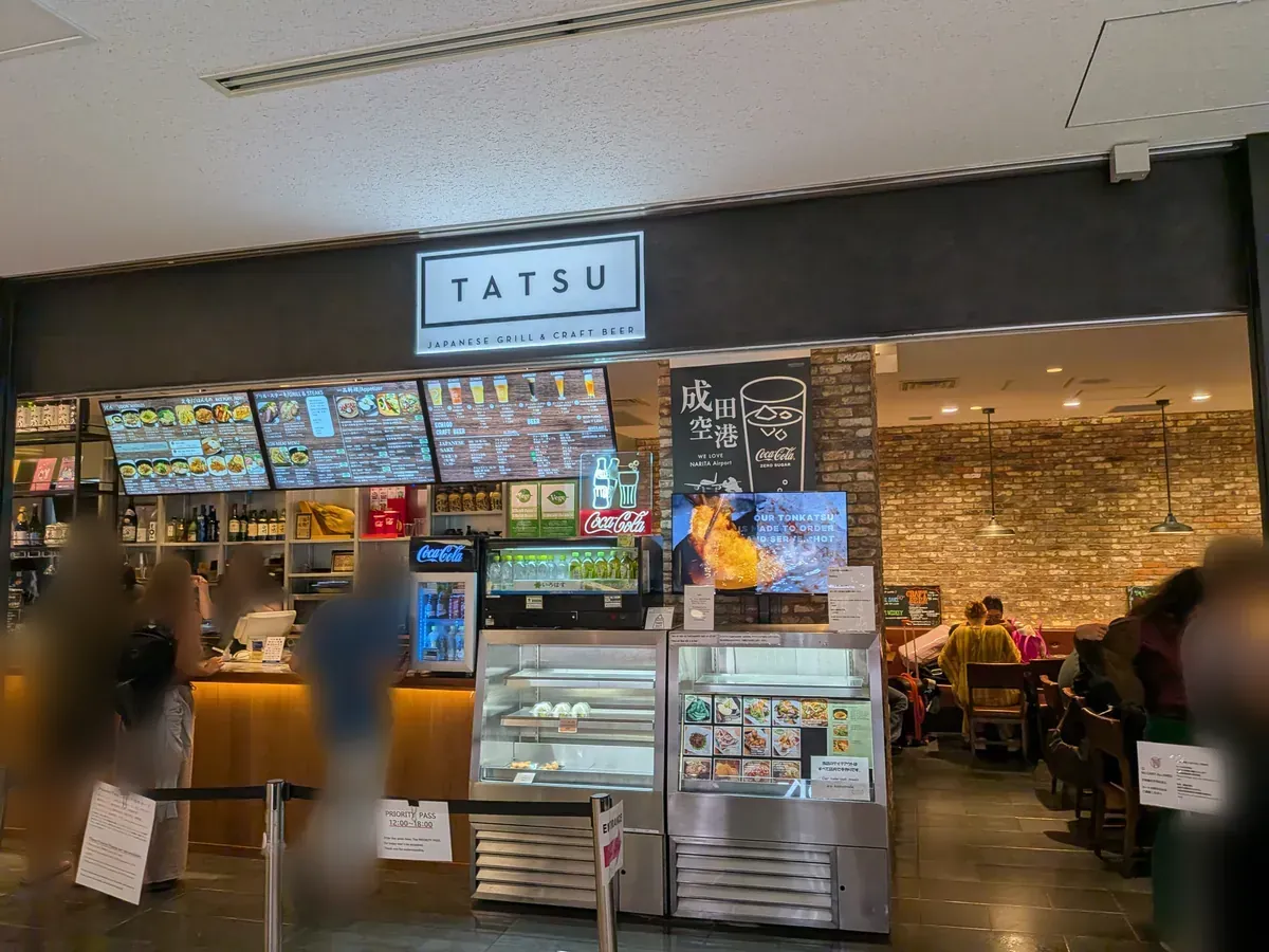 空港内のレストラン「TATSU JAPANESE GRILL & CRAFT BEER」の入口。メニューが掲示され、ショーケースには料理サンプルが並び、店内では食事が提供されている。