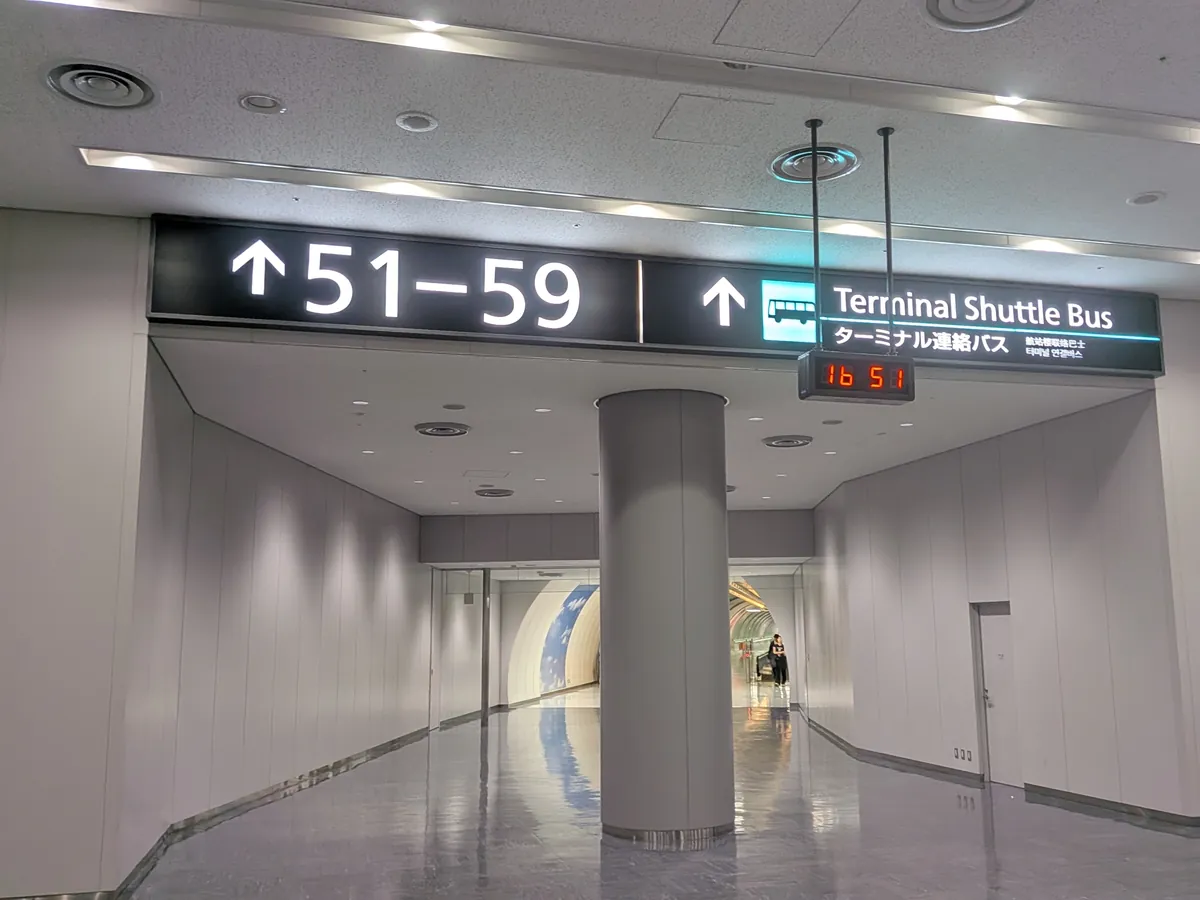 空港ターミナル内の通路に設置された案内表示板。「51-59番ゲート方面」と「ターミナル連絡バス」の方向が示され、シャトルバスの待ち時間も表示されている。