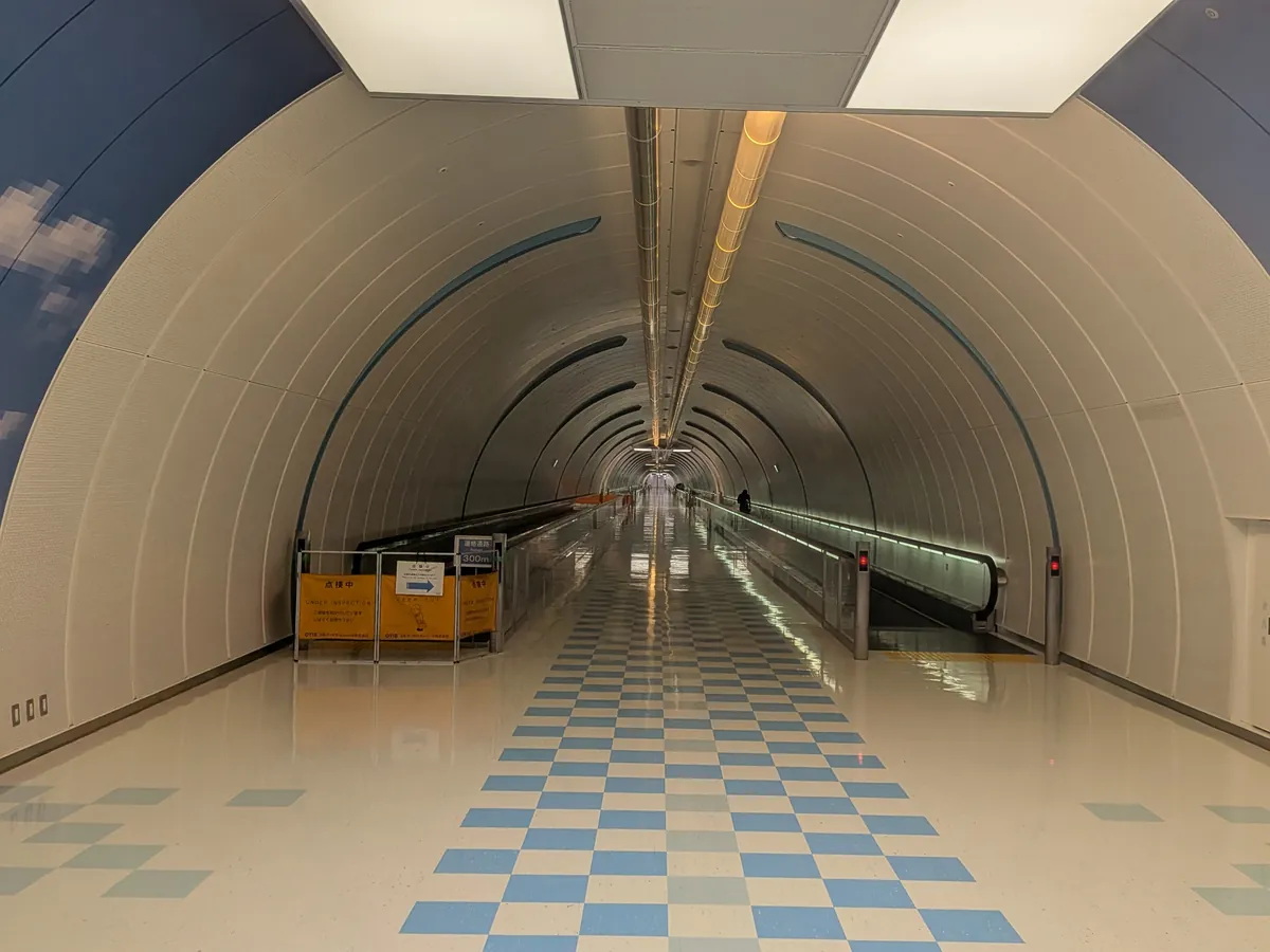 空港ターミナル内の、天井がアーチ状になった長い連絡通路。壁面には空の模様が描かれ、青と白の市松模様の床が続く動く歩道が設置されている。