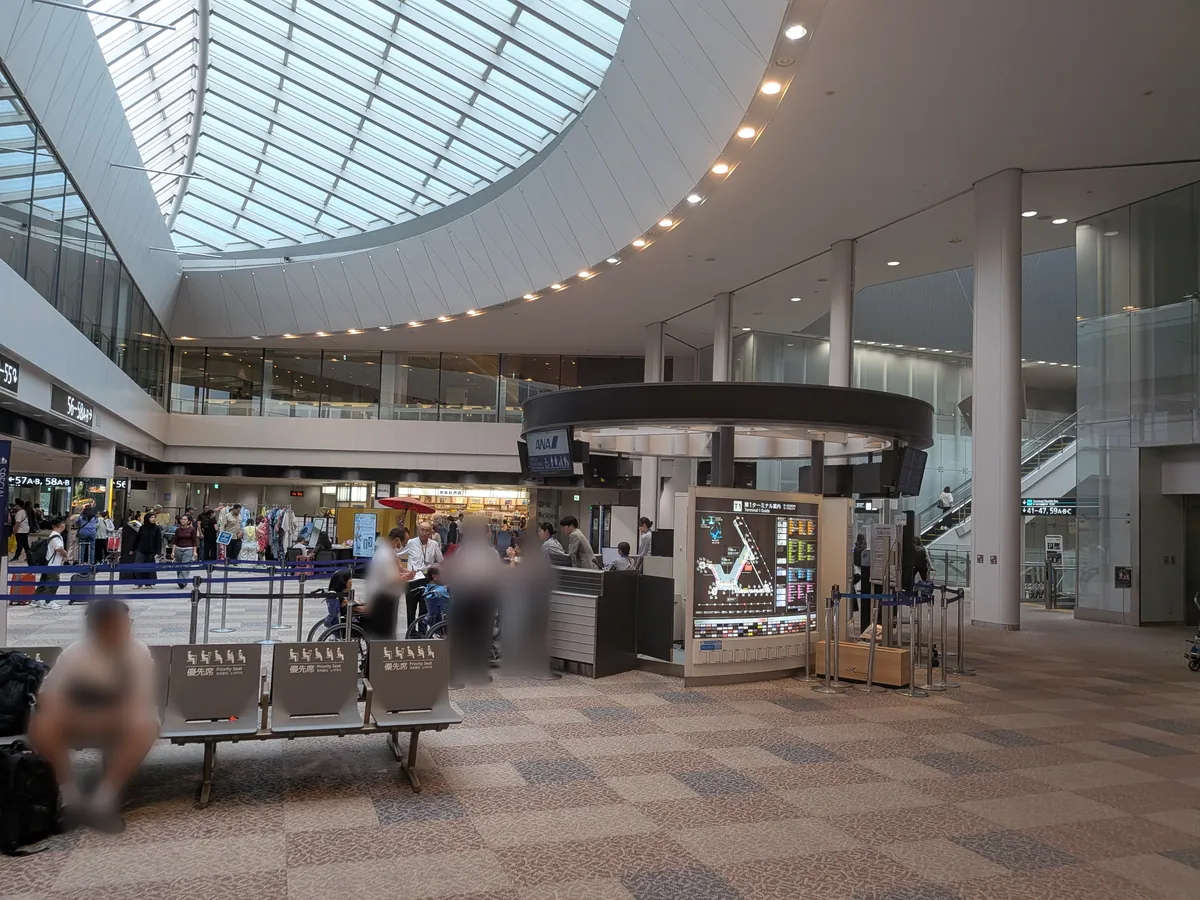 空港ターミナル内の広々とした待合エリア。大きなガラス張りの天井から自然光が差し込み、中央には案内カウンターが設置され、多くの人が行き交っている。
