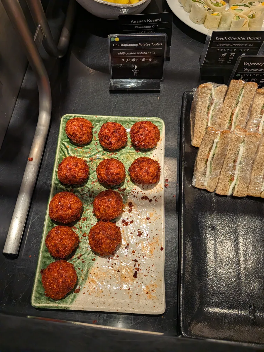 ビュッフェカウンターに並べられた、チリポテトボールとラップで個包装されたサンドイッチ。それぞれの料理名が英語と日本語で表示されている。