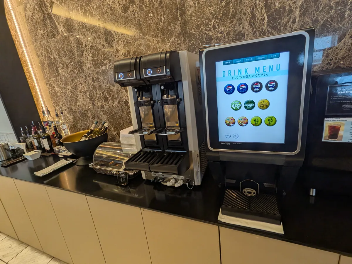 ラウンジ内のドリンクバー。ソフトドリンクのメニューが表示されたタッチパネル式の機械と、様々なアルコールボトルが並べられている。