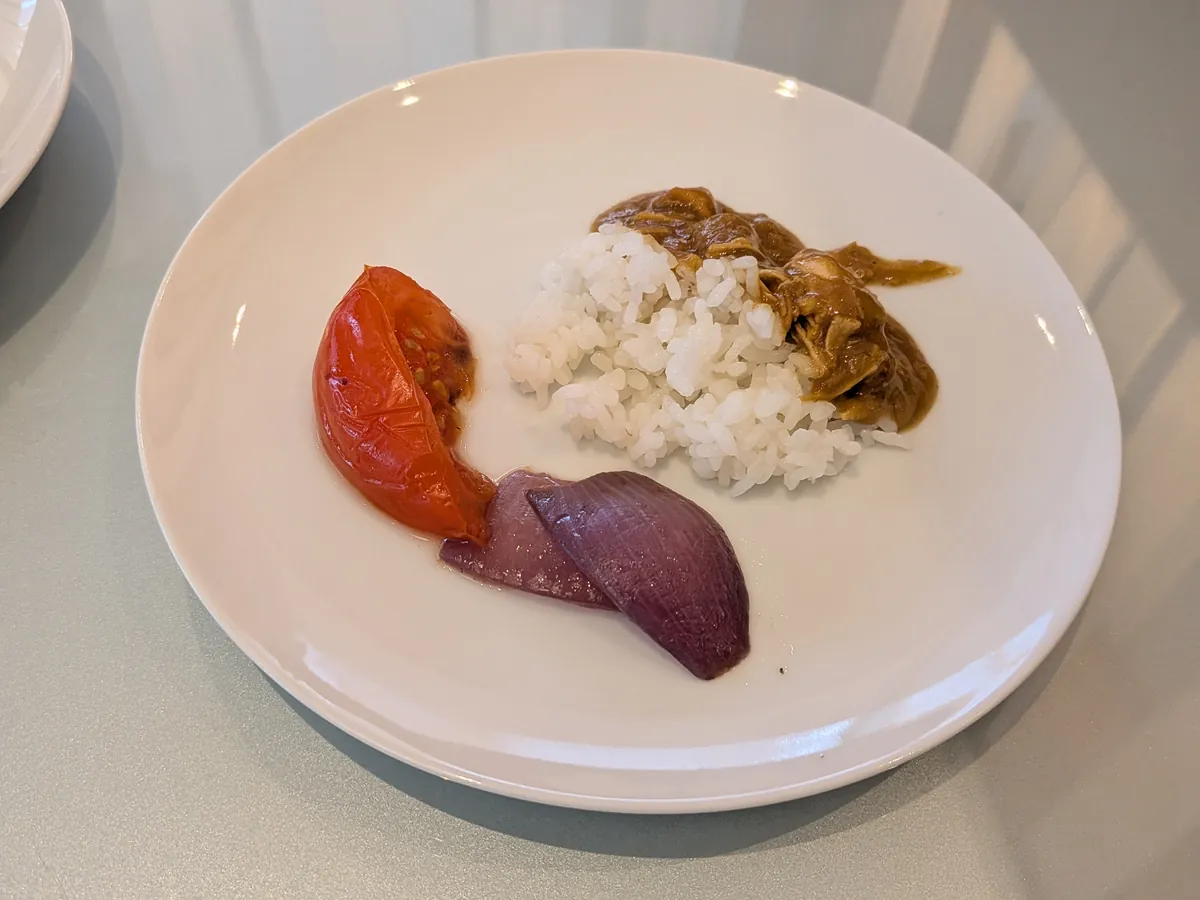 白い皿に盛られた食事。温かいトマトと赤玉ねぎ、白米、そしてカレーのようなソースが盛り付けられている。