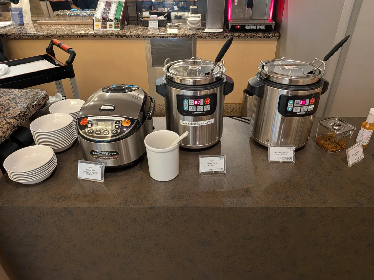 ラウンジのホットミールコーナー。ご飯ジャーとベジタリアンカレー、ポークカレーの保温器、取り皿が用意されている。