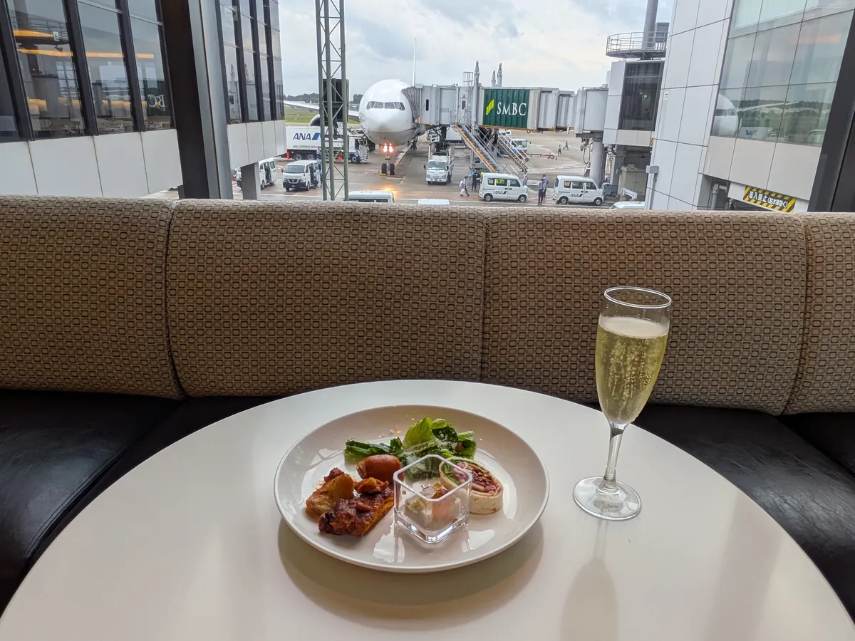 窓から飛行機が見えるラウンジ席で、軽食とスパークリングワインを楽しんでいる様子。