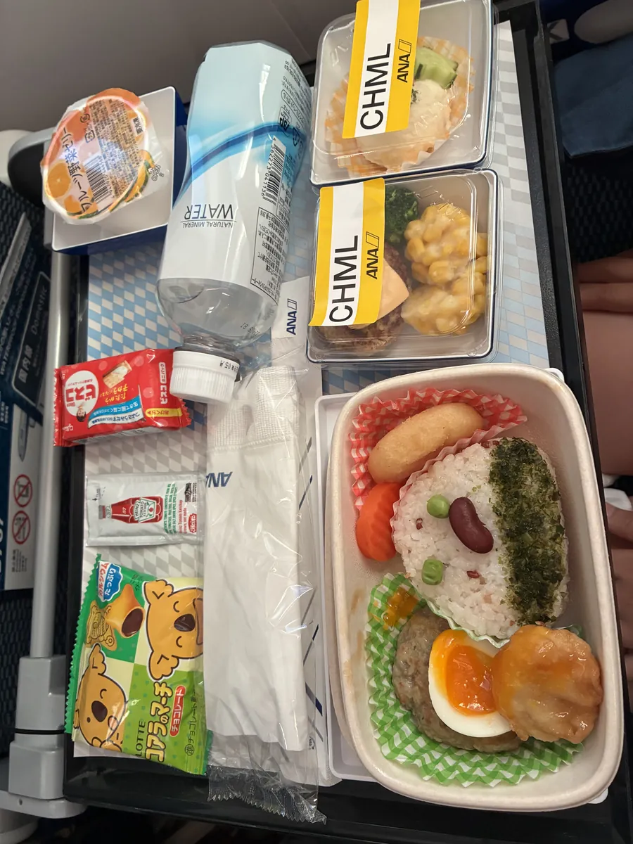 ANAのお子様向けのお食事(CHML)のトレイ。顔に見立てたおむすび、チーズハンバーグと半熟卵、から揚げ、ポテトサラダ、お菓子とゼリー
