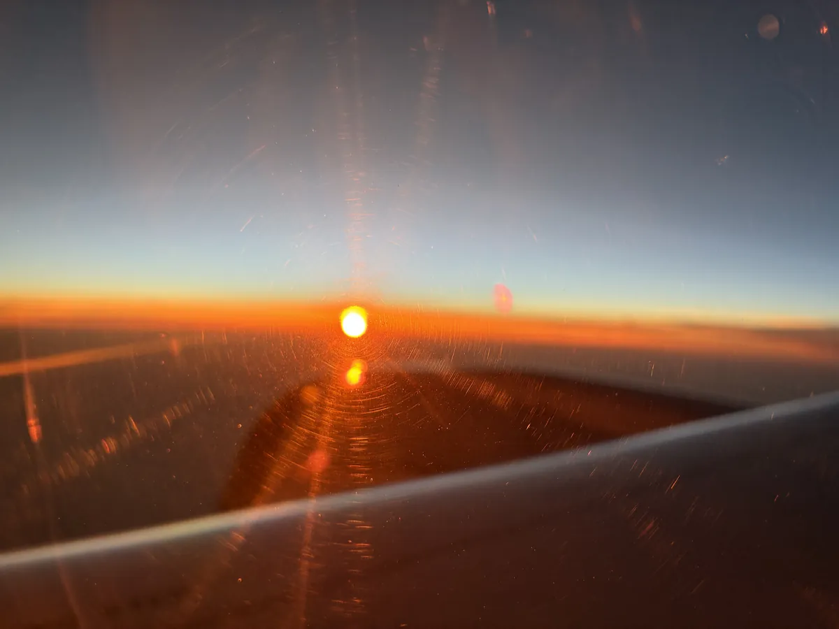 機内の窓から見た夕日。雲の上にオレンジ色の光が広がり、主翼の一部が見えている
