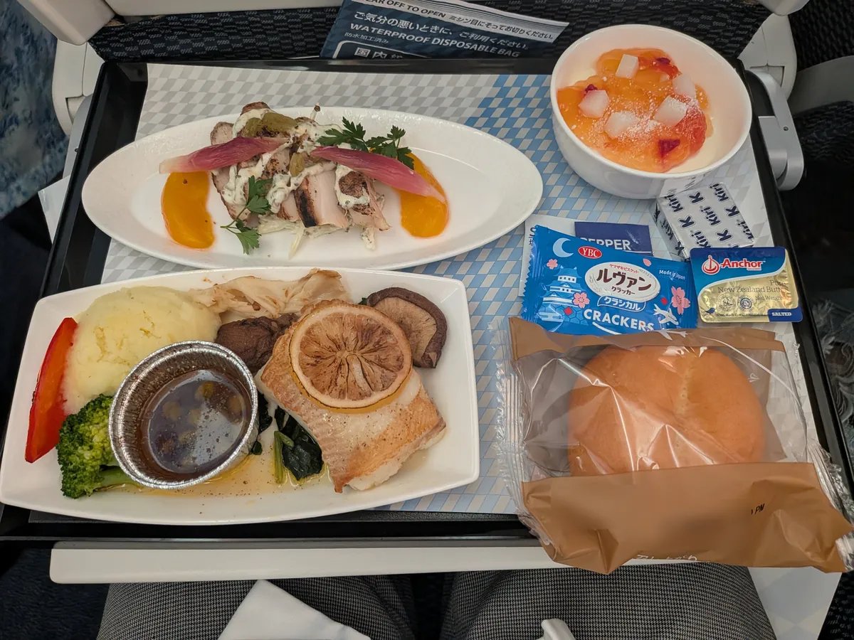 ANAの有料機内食(洋食)のトレイ。白い皿に盛られた前菜のスパイシーチキンと、レモンをのせたからす鰈のソテー、ムースライチとソフトロール