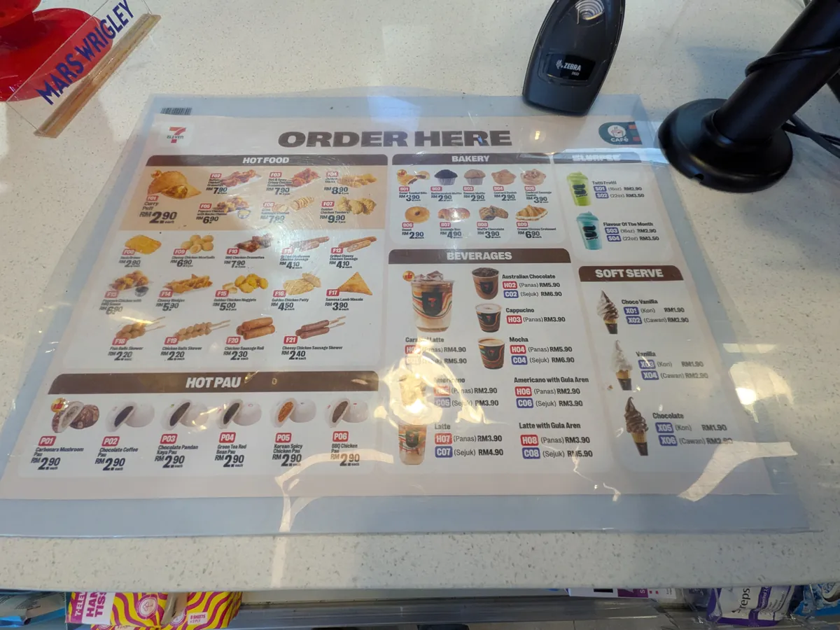 制限エリア内のコンビニのORDER HERE価格表。コーヒーやホットスナックの値段が写真つきで並ぶ