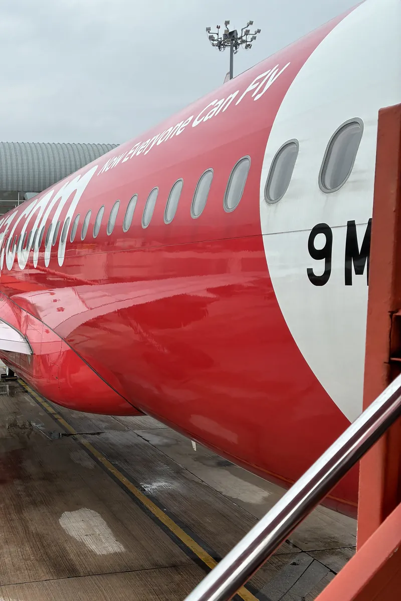 ペナン国際空港の駐機場に停まるエアアジアのエアバス。赤い機体に「Now Everyone Can Fly」の文字。雨上がりで濡れたエプロンとタラップの手すり
