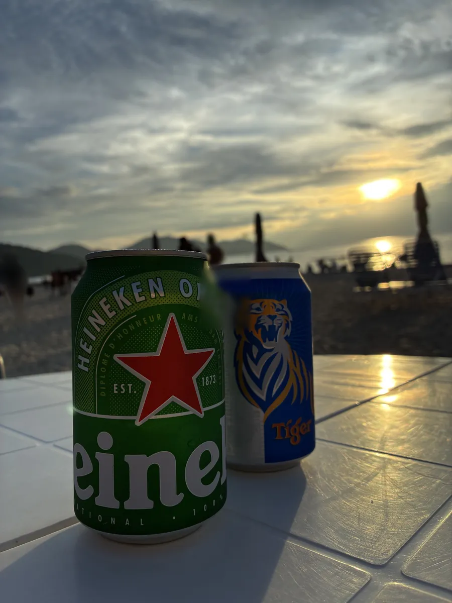 砂浜のテーブルに置かれた瓶ビールの缶と、その向こうに沈む夕日