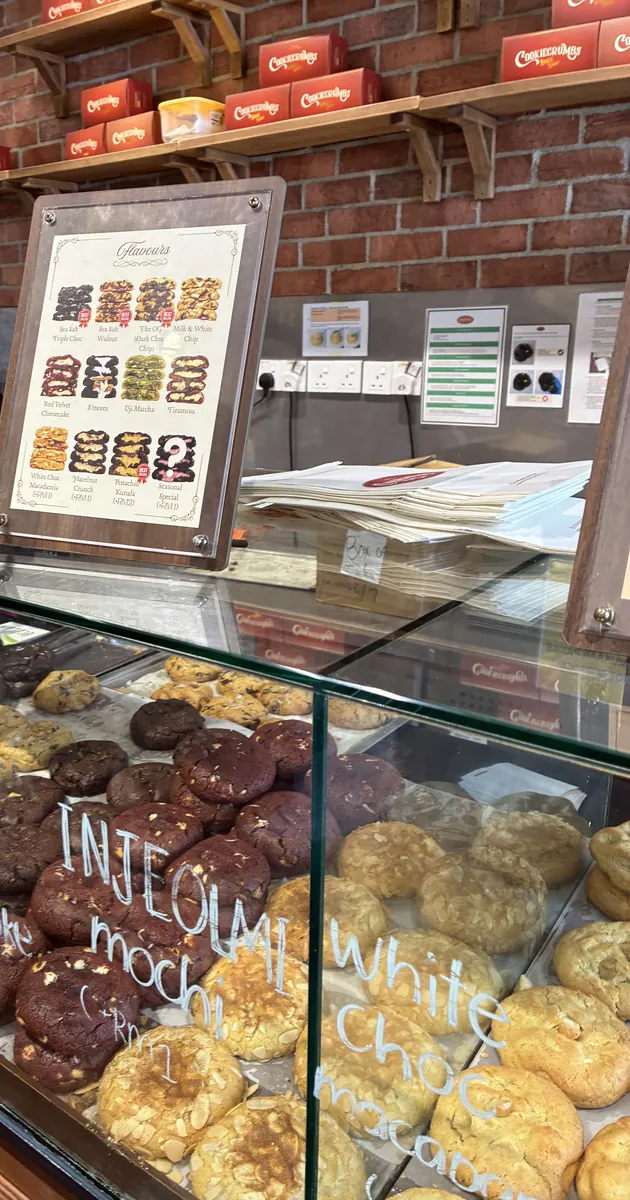 店内のショーケース。ガラスに白ペンでINJEOLMI mochiやWhite Choc Macadamiaと手書きされ、大ぶりのクッキーが並ぶ。奥のレンガ壁の棚には赤いギフトボックス