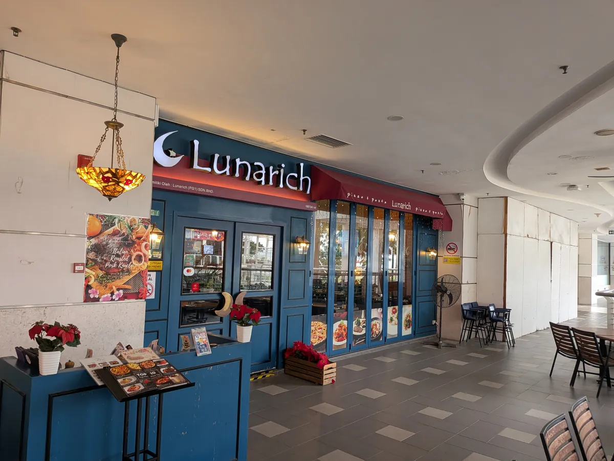 ショッピングモール2階のルナリッチ店舗外観。濃い青の木枠の扉と赤いひさし、白い『Lunarich』の看板。店頭に料理写真の立て看板とテラス席