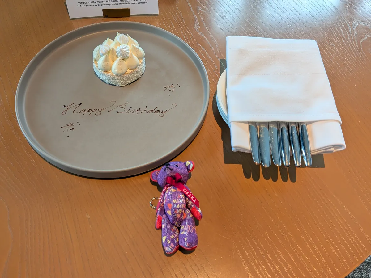 手書きでHappy Birthdayと描かれたプレートに載った小さなタルトと紫のコンラッドベア