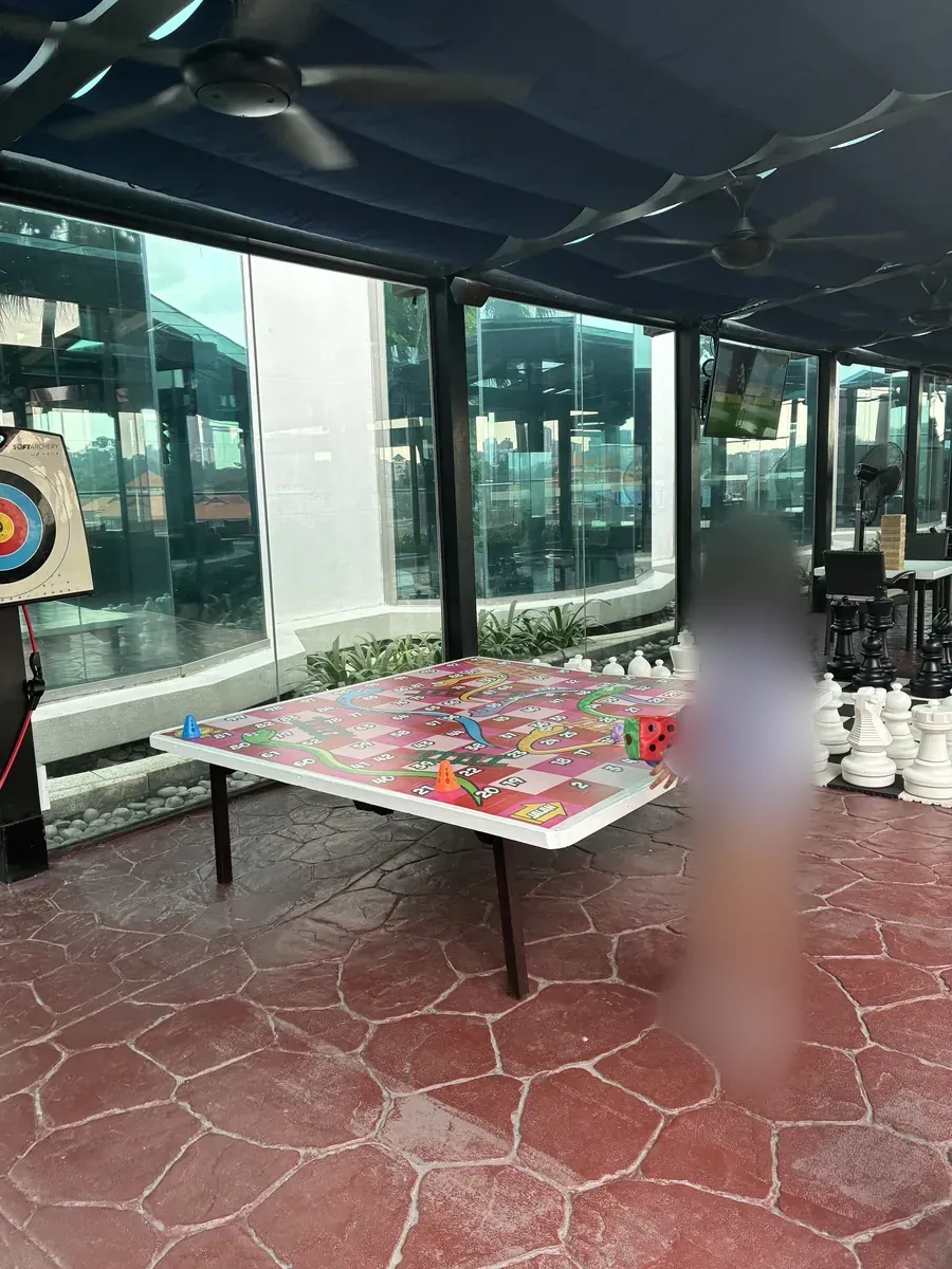 プールサイドの遊び場。卓球台の上に巨大な蛇と梯子のボードゲームが置かれ、奥に大きなチェスの駒とアーチェリーの的が並ぶ